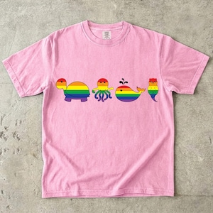 以下が含まれることがあります： 明るいピンク色の半袖Tシャツで、カメ、タコ、クジラ、カワウソの4種類の海の生き物が横一列に並んだイラストが描かれています。それぞれの生き物の体には、鮮やかな虹色のストライプ模様が施されています。