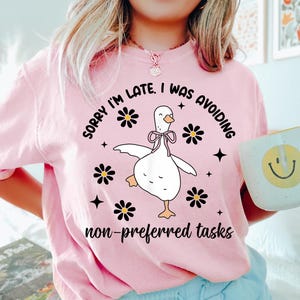Peut inclure: T-shirt rose avec l'inscription "SORRY I'M LATE, I WAS AVOIDING non-preferred tasks" et un dessin d'oie. L'oie est blanche avec un nœud rose, entourée de fleurs noires et d'étoiles. Une tasse blanche avec un smiley est tenue à la main.