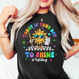 May include: Black t-shirt with a colorful graphic of a smiling sun wearing sunglasses, holding a pencil and a test paper. The text "TODAY IS YOUR DAY TO SHINE" and "#Testday" are also displayed.
