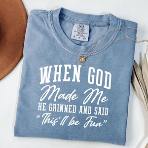May include: A blue Comfort Colors t-shirt with the text "WHEN GOD Made Me HE GRINNED AND SAID 'This'll be Fun'" in white. A gold necklace with a pendant is on the shirt.