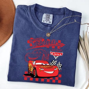 May include: A navy blue t-shirt with a graphic of Lightning McQueen from the Cars film. The design includes the text "Lightning McQueen EST 2006 Cars" in red, and checkered flags. A gold necklace with a pendant is draped across the shirt.