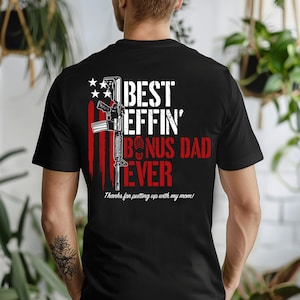 May include: A black t-shirt featuring a graphic on the back with a white rifle silhouette overlaid on red and white vertical stripes. The text reads BEST EFFIN' BONUS DAD EVER in white and red, with the phrase Thanks for putting up with my mom!
