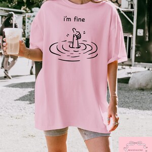 May include: Pink oversized t-shirt with a black graphic of a hand giving a thumbs up while submerged in water. The text "i'm fine" is printed above the graphic.
