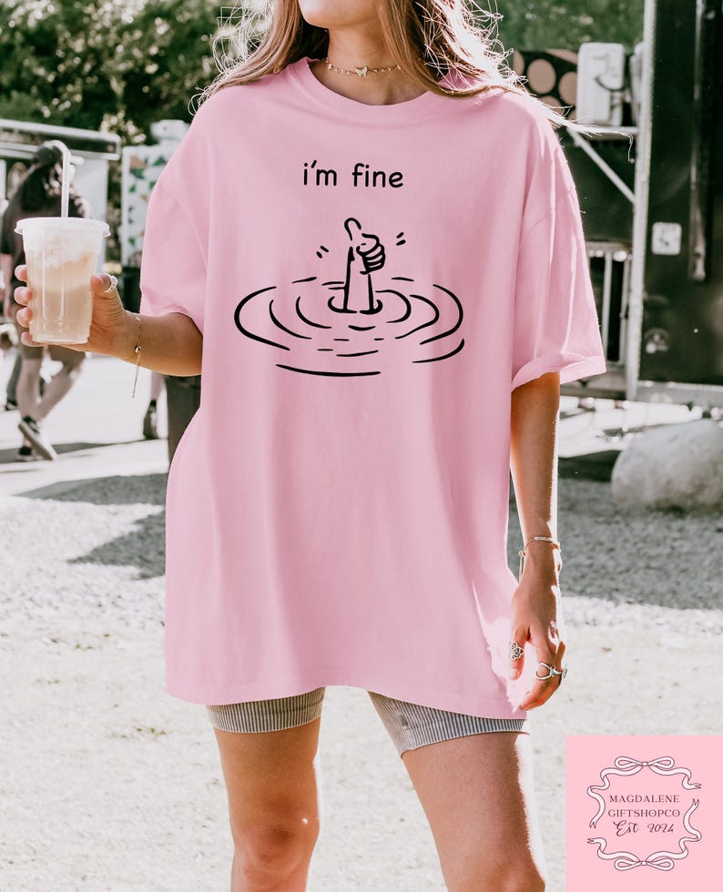 May include: Pink oversized t-shirt with a black graphic of a hand giving a thumbs up while submerged in water. The text "i'm fine" is printed above the graphic.