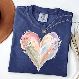 May include: A navy blue short-sleeve t-shirt featuring a large, painterly heart graphic in soft pastel shades of pink, peach, and lavender, accented with delicate floral illustrations. The inner neck label reads Comfort Colors, 100% cotton, S-P-CH.