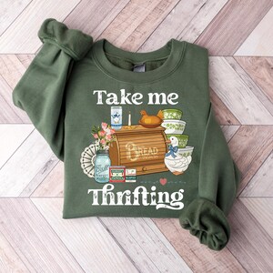 May include: Green sweatshirt with the words "Take me Thrifting" and a graphic of a bread box, bowls, and a duck. The sweatshirt is a solid green color with a crew neck and long sleeves.