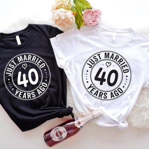 May include: Two black and white t-shirts with the text "Just Married 40 Years Ago" in a circle with a heart. The shirts are laid out on a white surface.
