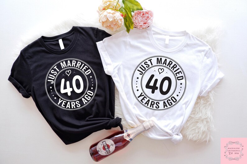 May include: Two black and white t-shirts with the text "Just Married 40 Years Ago" in a circle with a heart. The shirts are laid out on a white surface.