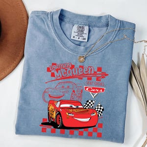 May include: A light blue t-shirt with a graphic of Lightning McQueen from the Cars film. The design includes the text "Lightning McQueen" and "Cars Est 2006". The shirt has a crew neck and short sleeves.