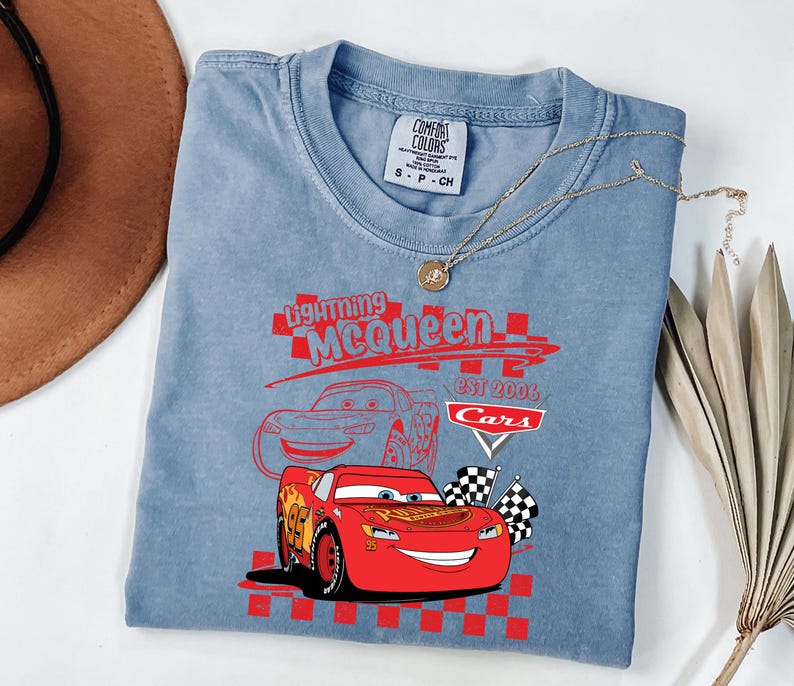 May include: A light blue t-shirt with a graphic of Lightning McQueen from the Cars film. The design includes the text "Lightning McQueen" and "Cars Est 2006". The shirt has a crew neck and short sleeves.