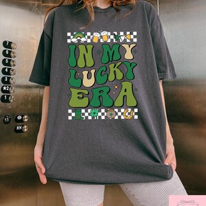 Puede incluir: Una camiseta gris oscuro con un patr&oacute;n a cuadros y el texto "IN MY LUCKY ERA" en letras verdes y amarillas. El dise&ntilde;o incluye un arco&iacute;ris, jarras de cerveza, tr&eacute;boles, una olla de oro y una herradura.