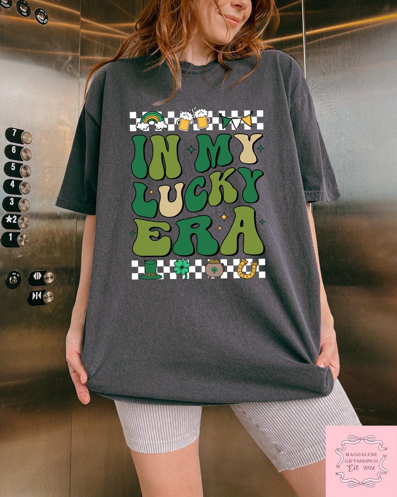 Puede incluir: Una camiseta gris oscuro con un patr&oacute;n a cuadros y el texto "IN MY LUCKY ERA" en letras verdes y amarillas. El dise&ntilde;o incluye un arco&iacute;ris, jarras de cerveza, tr&eacute;boles, una olla de oro y una herradura.