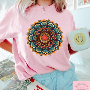 May include: A light pink t-shirt featuring a vibrant, detailed mandala design in shades of blue, yellow, red, and orange. The shirt is paired with light blue shorts. A white mug with a yellow smiley face is held in the hand.