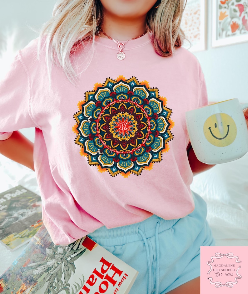 May include: A light pink t-shirt featuring a vibrant, detailed mandala design in shades of blue, yellow, red, and orange. The shirt is paired with light blue shorts. A white mug with a yellow smiley face is held in the hand.