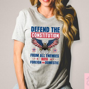 May include: A light grey t-shirt with a patriotic design. The design features the text "DEFEND THE CONSTITUTION" above an eagle illustration with American flag-themed wings. Below the eagle, the text reads "FROM ALL ENEMIES BOTH FOREIGN AND DOMESTIC."