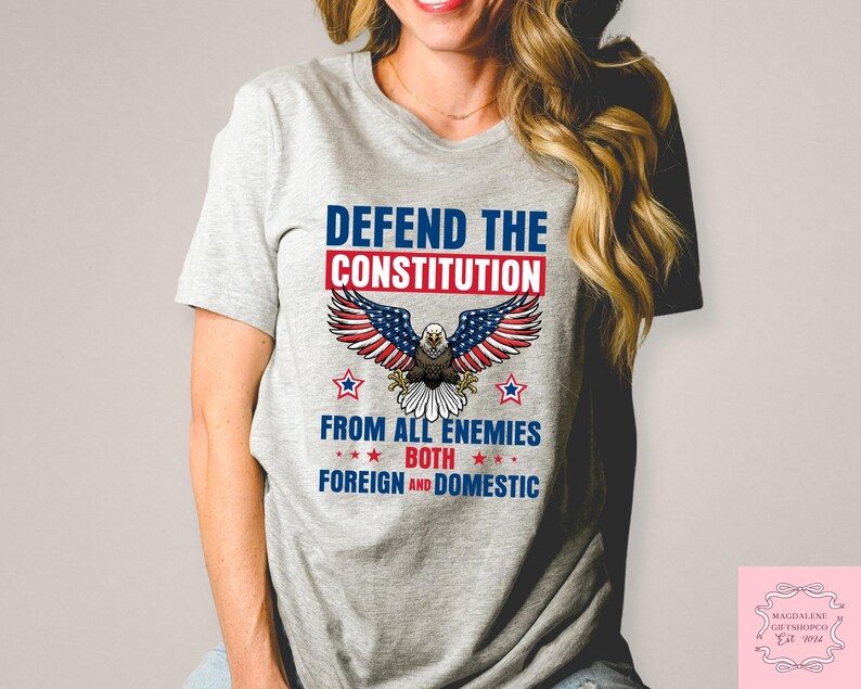 May include: A light grey t-shirt with a patriotic design. The design features the text "DEFEND THE CONSTITUTION" above an eagle illustration with American flag-themed wings. Below the eagle, the text reads "FROM ALL ENEMIES BOTH FOREIGN AND DOMESTIC."