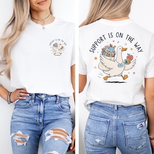 Op de afbeelding: Een wit t-shirt met een afbeelding van een eend die schoolspullen en een appel draagt. De voorkant toont een kleine eend met de tekst "SUPPORT IS ON THE WAY", terwijl de achterkant een grotere versie van dezelfde eend laat zien.