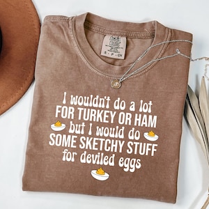 May include: A brown Comfort Colors t-shirt with the text "I wouldn't do a lot FOR TURKEY OR HAM but I would do SOME SKETCHY STUFF for deviled eggs." A silver necklace with a pendant is on the shirt.