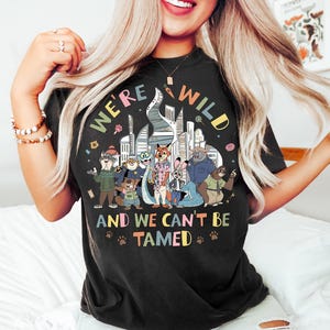 May include: Black t-shirt with a colorful graphic featuring cartoon animals in front of a city skyline. The text on the shirt reads "WE'RE WILD" and "AND WE CAN'T BE TAMED" with paw prints.