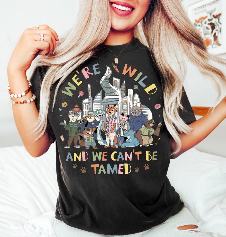 May include: Black t-shirt with a colorful graphic featuring cartoon animals in front of a city skyline. The text on the shirt reads "WE'RE WILD" and "AND WE CAN'T BE TAMED" with paw prints.