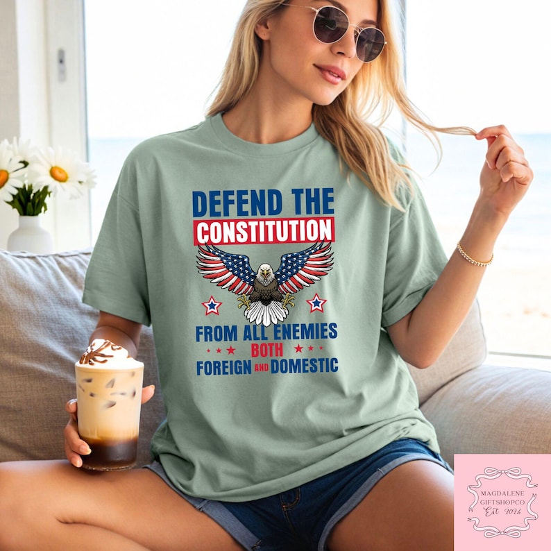 May include: A pale green t-shirt featuring the words "DEFEND THE CONSTITUTION" above an eagle design with red, white, and blue wings. The text "FROM ALL ENEMIES BOTH FOREIGN AND DOMESTIC" is below the eagle. The shirt is worn by a person.
