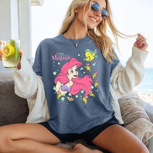 Puede incluir: Camiseta azul con un gr&aacute;fico de Ariel de La Sirenita de Disney. Ariel tiene el pelo rosa y un top de concha p&uacute;rpura. La camiseta tambi&eacute;n presenta el texto "Disney The Little Mermaid" e im&aacute;genes de Flounder y caballitos de mar.