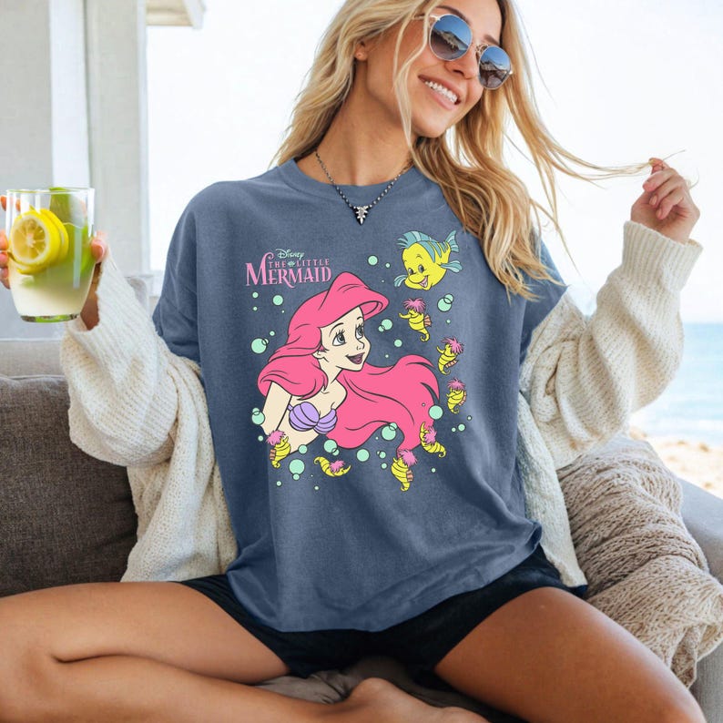 Puede incluir: Camiseta azul con un gr&aacute;fico de Ariel de La Sirenita de Disney. Ariel tiene el pelo rosa y un top de concha p&uacute;rpura. La camiseta tambi&eacute;n presenta el texto "Disney The Little Mermaid" e im&aacute;genes de Flounder y caballitos de mar.