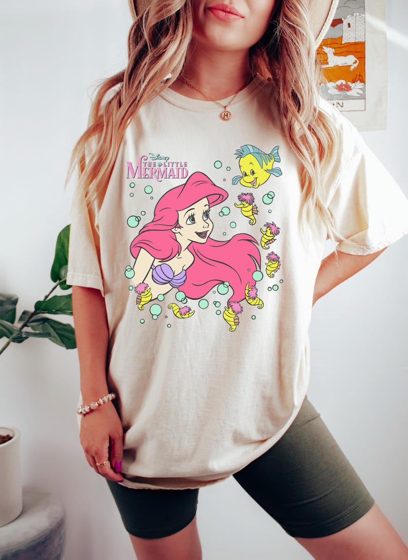 Puede incluir: Camiseta color crema con un gr&aacute;fico de Ariel de La Sirenita de Disney. Ariel tiene el pelo rosa y un top de concha p&uacute;rpura. La camiseta tambi&eacute;n presenta a Flounder y otras criaturas marinas. El texto "Disney The Little Mermaid" est&aacute; arriba.