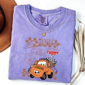May include: A lilac t-shirt with a graphic of the character Tow Mater from the film Cars. The design includes the text "Tow Matter" and "Cars Est 2006". A gold necklace and a brown hat are also visible.