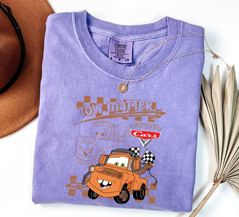 May include: A lilac t-shirt with a graphic of the character Tow Mater from the film Cars. The design includes the text "Tow Matter" and "Cars Est 2006". A gold necklace and a brown hat are also visible.
