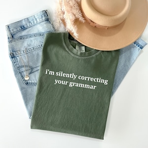 May include: An olive green t-shirt with the text "I'm silently correcting your grammar" in white. The shirt is laid on light blue jeans, with a gold necklace and a tan hat.