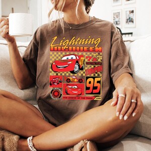 May include: Brown t-shirt with a graphic of Lightning McQueen from the Cars film. The design includes the text "Lightning McQueen" in yellow, the number "95", and images of the character. The shirt has a relaxed fit.