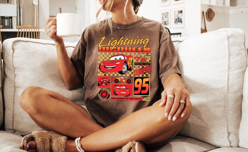 May include: Brown t-shirt with a graphic of Lightning McQueen from the Cars film. The design includes the text "Lightning McQueen" in yellow, the number "95", and images of the character. The shirt has a relaxed fit.