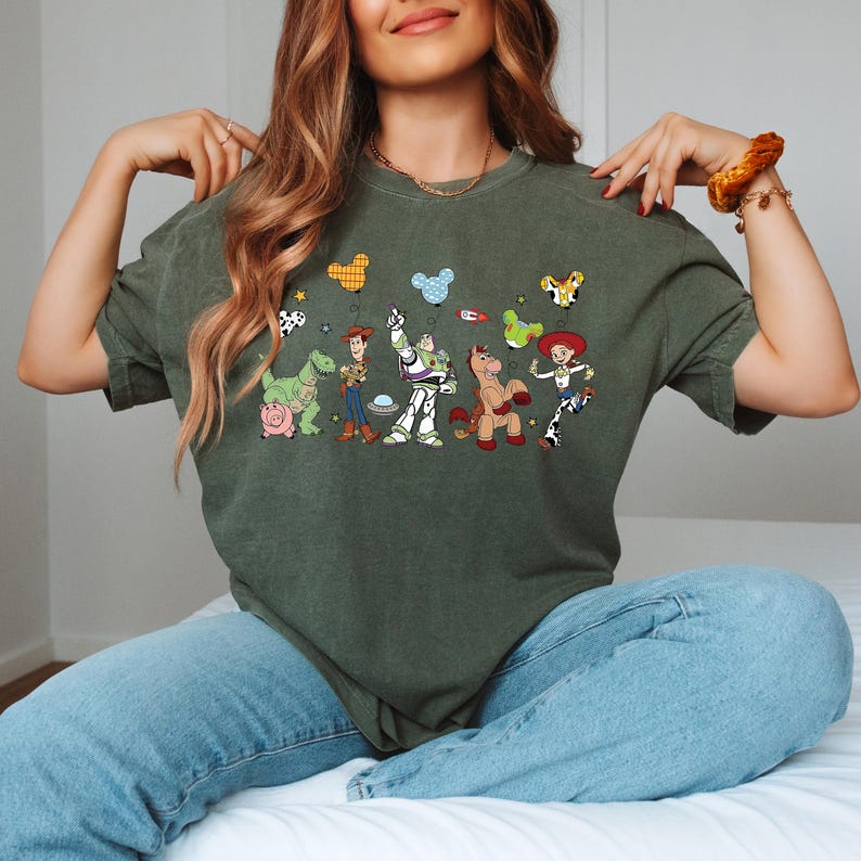 May include: An olive green t-shirt featuring a colorful graphic of animated characters. The design includes a dinosaur, a cowboy, a space ranger, a horse, and a cowgirl. The shirt is made of a soft material and has short sleeves.