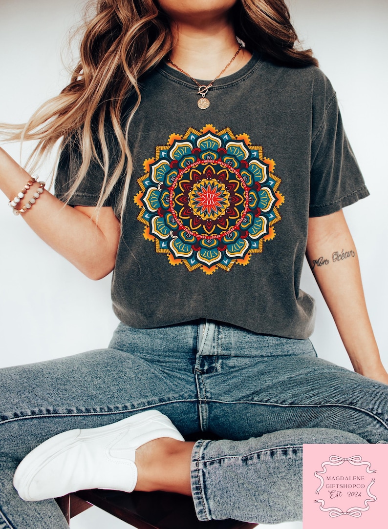 May include: Dark gray t-shirt featuring a vibrant, colorful mandala design. The mandala is a circular pattern with shades of red, yellow, blue, and green. The shirt is paired with blue jeans and white sneakers. The image also includes a necklace and bracelets.