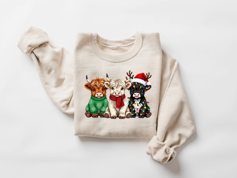 Pu&ograve; includere: Una felpa color crema con una grafica di tre mucche delle Highlands. Una mucca indossa un maglione verde, un'altra una sciarpa rossa e la terza un cappello di Babbo Natale e luci natalizie. Le mucche sono sedute, creando un tema festivo.