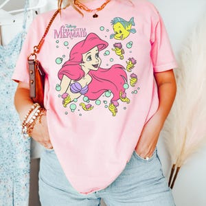 Puede incluir: Camiseta rosa claro con un dise&ntilde;o de Ariel de La Sirenita de Disney. El dise&ntilde;o incluye a Ariel con pelo rosa, un sujetador de concha p&uacute;rpura y personajes de peces y caballitos de mar amarillos. La camiseta tiene el texto "Disney The Little Mermaid".