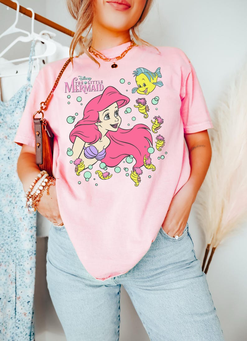 Puede incluir: Camiseta rosa claro con un dise&ntilde;o de Ariel de La Sirenita de Disney. El dise&ntilde;o incluye a Ariel con pelo rosa, un sujetador de concha p&uacute;rpura y personajes de peces y caballitos de mar amarillos. La camiseta tiene el texto "Disney The Little Mermaid".