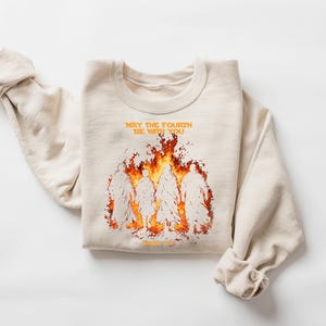 K&ouml;nnte beinhalten: Beiges Sweatshirt mit Rundhalsausschnitt und dem orangefarbenen Text "MAY THE FOURTH BE WITH YOU". Darunter eine Grafik von Figuren in Flammen. Die &Auml;rmel sind hochgekrempelt. Das Sweatshirt ist gefaltet.