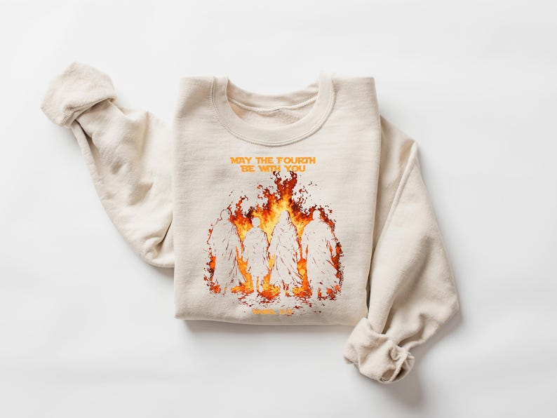 K&ouml;nnte beinhalten: Beiges Sweatshirt mit Rundhalsausschnitt und dem orangefarbenen Text "MAY THE FOURTH BE WITH YOU". Darunter eine Grafik von Figuren in Flammen. Die &Auml;rmel sind hochgekrempelt. Das Sweatshirt ist gefaltet.
