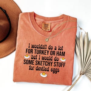 May include: A coral-colored t-shirt with the text "I wouldn't do a lot for turkey or ham but I would do some sketchy stuff for deviled eggs." A gold necklace and a brown hat are also in the image.