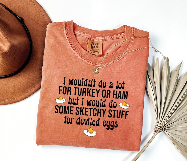 May include: A coral-colored t-shirt with the text "I wouldn't do a lot for turkey or ham but I would do some sketchy stuff for deviled eggs." A gold necklace and a brown hat are also in the image.