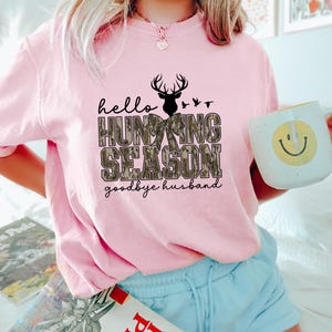 K&ouml;nnte beinhalten: Hellrosa Sweatshirt mit dem Text "hello HUNTING SEASON goodbye husband" in Schwarz und Camouflage-Print. Das Design beinhaltet eine Hirschsilhouette und V&ouml;gel. Die Person h&auml;lt einen wei&szlig;en Becher mit einem gelben Smiley.