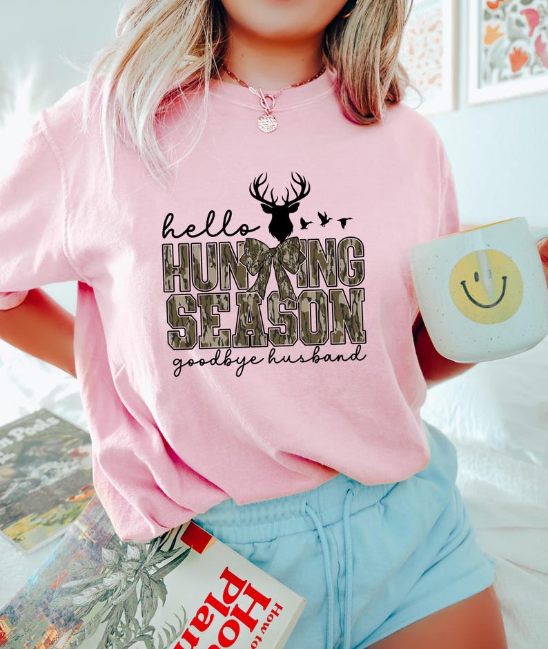 K&ouml;nnte beinhalten: Hellrosa Sweatshirt mit dem Text "hello HUNTING SEASON goodbye husband" in Schwarz und Camouflage-Print. Das Design beinhaltet eine Hirschsilhouette und V&ouml;gel. Die Person h&auml;lt einen wei&szlig;en Becher mit einem gelben Smiley.