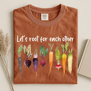 Può includere: Una t-shirt color ruggine con il testo bianco "Let's root for each other" sopra una colorata illustrazione ad acquerello di vari ortaggi a radice, tra cui carote, barbabietole e ravanelli con foglie verdi.