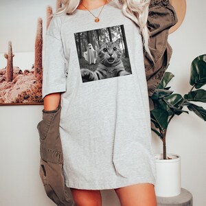 May include: A light gray t-shirt dress featuring a black and white image of a cat taking a selfie with a ghost in a forest. The cat has wide eyes and an open mouth. The dress is a loose fit.