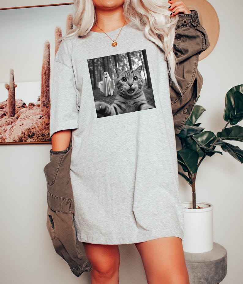 May include: A light gray t-shirt dress featuring a black and white image of a cat taking a selfie with a ghost in a forest. The cat has wide eyes and an open mouth. The dress is a loose fit.