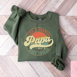 May include: An olive green sweatshirt with the text "Like a Grandpa, Papa Only Cooler" in a retro design. The graphic features a circular design with the word "Papa" in a large, stylized font, with a sunset-like background.