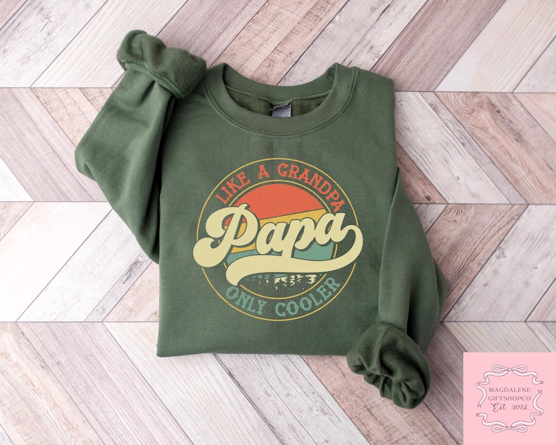 May include: An olive green sweatshirt with the text "Like a Grandpa, Papa Only Cooler" in a retro design. The graphic features a circular design with the word "Papa" in a large, stylized font, with a sunset-like background.