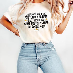 May include: Cream-colored t-shirt with the text "I wouldn't do a lot FOR TURKEY OR HAM but I would do SOME SKETCHY STUFF for deviled eggs." The shirt is paired with light blue jeans. The text is in black.